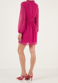 Fuchsia chiffongklänning med långa ärmar, smockad midja och volang i halsringningen, kombinerad med högklackade skor i starkt rosa.