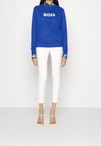 Modrá mikina s textem "BOSS", kombinovaná s bílými úzkými kalhotami a průhlednými sandály. Mezi výrazné prvky patří kontrastní vzorované manžety límce.