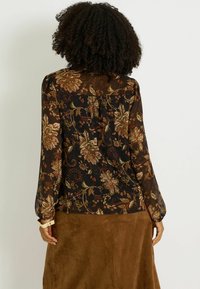Blusa negra transparente con un patrón floral en verde, óxido y crema. Mangas largas, hombros ligeramente fruncidos y un dobladillo redondeado. Falda marrón visible.