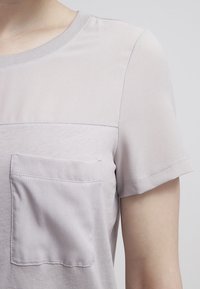 Světle šedá košile s krátkým rukávem, oblé výstřihem, přední kapsou a směsí hladké a mírně strukturované látky.