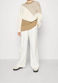 Osoba v béžovo-krémovém pleteném svetr s barevnými bloky, bílých kalhotách s širokými nohavicemi a světle béžových botách s černými podrážkami.