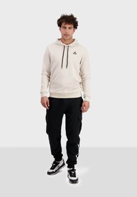 Sudadera de felpa crema con un bolsillo frontal y cordones negros, combinada con pantalones jogger negros que presentan bolsillos cargo y detalles de logo.