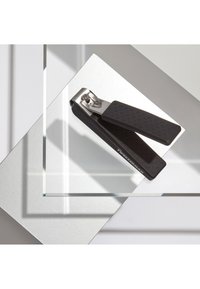 Zwarte Tweezerman nagelknipper met een gestructureerde grip, rustend op gelaagde glas- en zilveroppervlakken die geometrische schaduwen werpen.