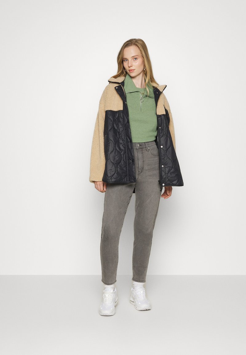 Chaqueta acolchada negra y beige con cuello alto, camiseta polo verde, jeans grises y zapatillas blancas; atuendo casual en capas.