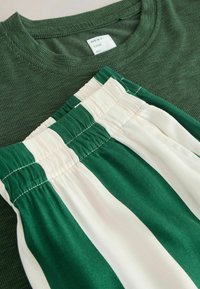 Grünes geripptes T-Shirt mit Rundhalsausschnitt über einer grünen und cremefarbenen gestreiften Shorts mit elastischem Bund, die aus glattem Stoff mit geraffter Textur gefertigt sind.