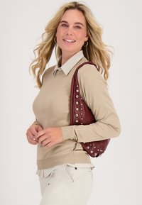 Beige langmouwen top met een lichte kraag, gecombineerd met beige broek. Houdt een bordeauxrode tas vast, versierd met zilveren studs. Gladde stofstructuur.