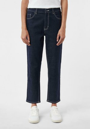 Personne portant un jean droit bleu foncé avec des coutures blanches et des baskets blanches, debout devant un fond uni.