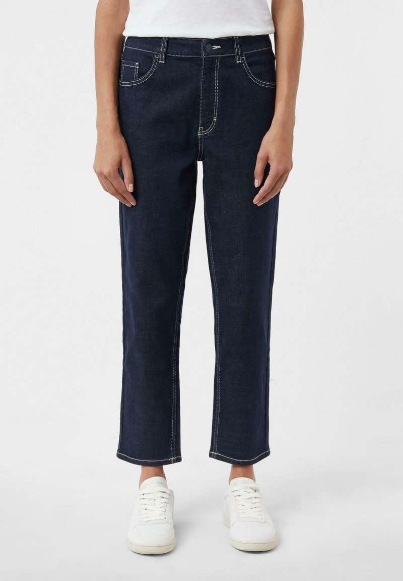 Personne portant un jean droit bleu foncé avec des coutures blanches et des baskets blanches, debout devant un fond uni.