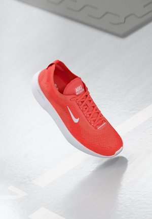 Lyse rød Nike løbesko med hvid swoosh-logo og hvid sål, vist svævende over en lysegrå overflade med svage linjer.