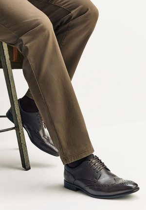 Homme portant un pantalon marron habillé et des chaussures Oxford en cuir brun foncé à bouts ailés, assis sur un tabouret en métal.