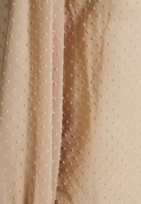 Tissu beige avec une surface texturée présentant de petits points surélevés, créant un motif subtil. Matériau doux et léger avec une finition lisse.