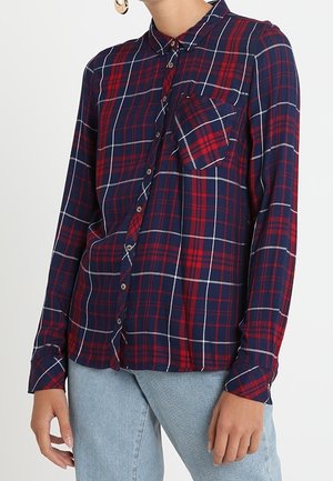 Vrouw draagt een lange mouwen blauw-rood geruit overhemd met borstzak, gecombineerd met lichtblauwe jeans.