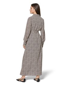 Lang, bruin en crèmekleurig jurk met luipaardprint, met een kraag, lange mouwen, een ceintuur in de taille en een zwierige rok. Draagt zwarte platte schoenen.