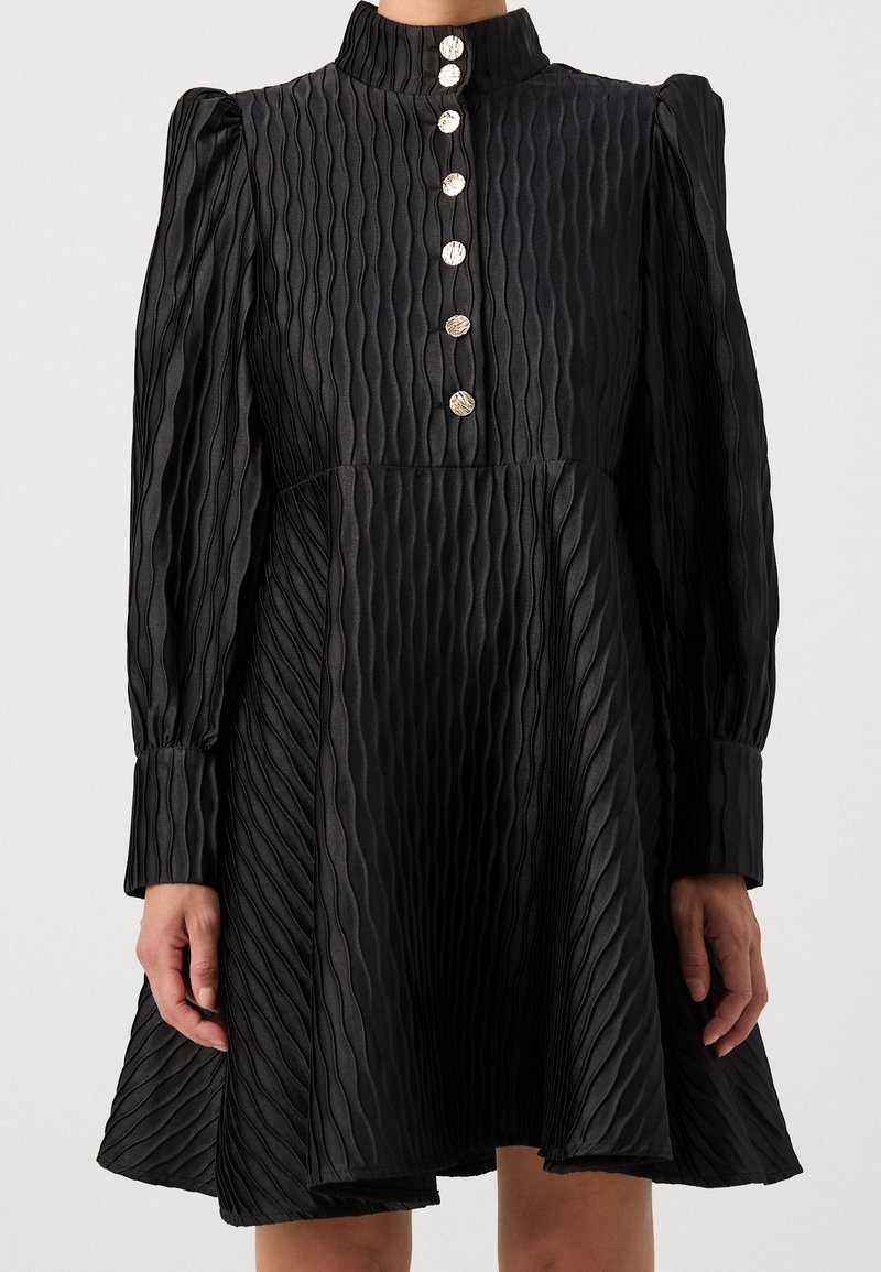 Robe longue noire texturée à manches longues avec col montant, boutons dorés sur le devant et jupe évasée, portée par une personne montrée des épaules aux genoux.