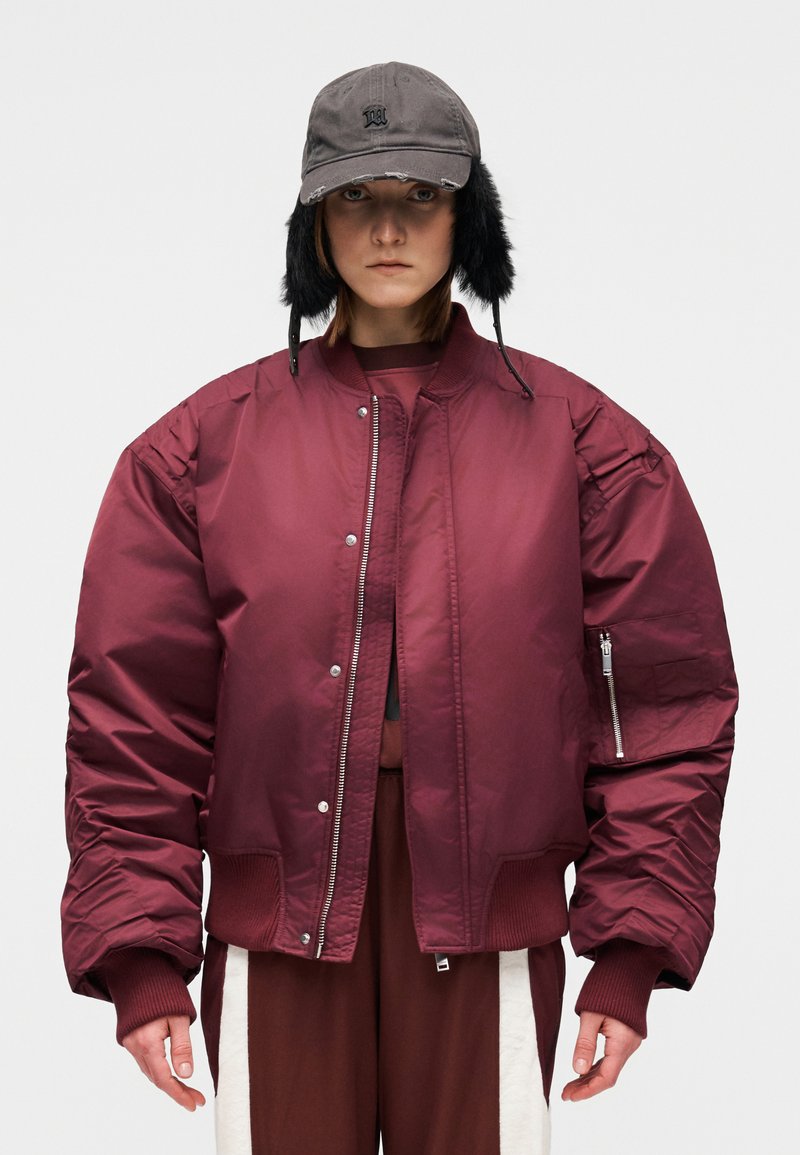 Veste bomber bordeaux à texture matelassée, manches avec poignets côtelés, et poche à fermeture éclair. Associée à une casquette grise avec cache-oreilles noirs.