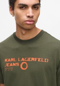 Mladý muž s kudrnatými vlasy nosí olivově zelené tričko s oranžovým nápisem "Karl Lagerfeld Jeans" a malou kulatou grafikou na přední straně.