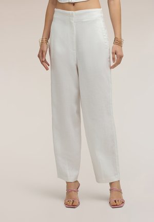Femme portant un pantalon blanc taille haute à jambes larges et des talons roses à brides, debout devant un fond neutre et uni.