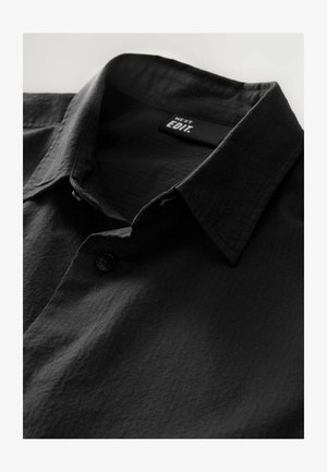 Cămașă neagră cu guler button-down, țesătură texturată cu un model subtil și etichetă care spune "NEXT EDIT." Finisaj neted, mat.