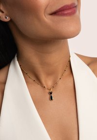 Collar de oro con un colgante negro en forma de gato con cola. La cadena es delicada y tiene pequeñas cuentas negras intercaladas.