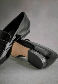 Black patent leather loafers with a rounded toe, textured rubber sole, and flat heel. Features a smooth surface and subtle stitching detail.