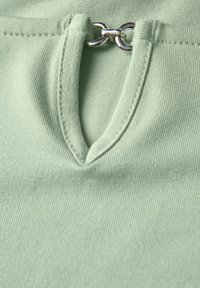 Světlý zelený top s texturovanou látkou, kulatým výstřihem a malým klíčovým výřezem ozdobeným stříbrným kovovým akcentem.