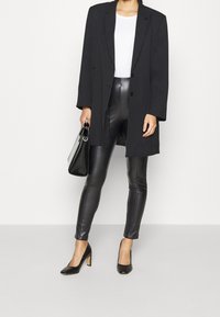 Černý oversized blazer, bílý tričko, černé legíny z umělé kůže, černé špičaté podpatky a černá kabelka, vše v minimalistickém stylu.