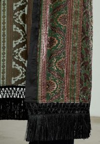 Tkanină decorativă cu modele florale verzi, maronii și aurii. Are o margine franjurată neagră și un design în dungi de lățimi variate.