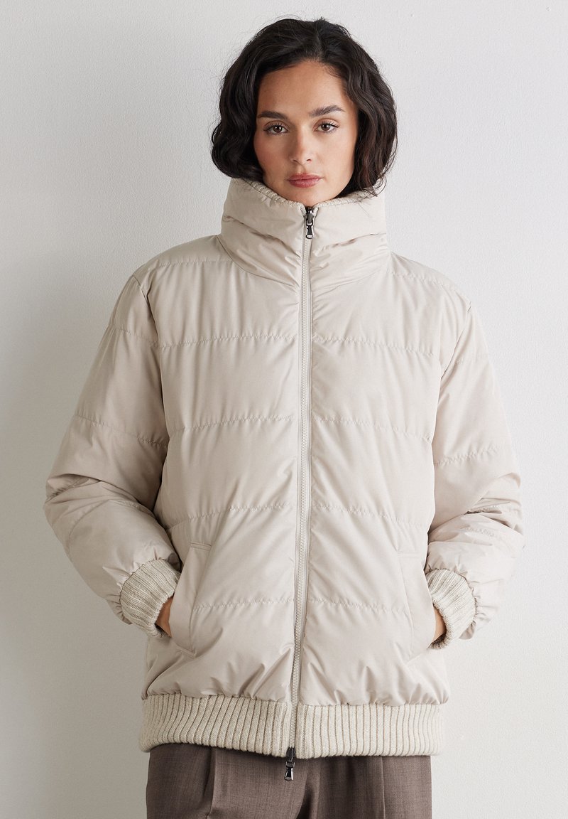 Kvinde iført en beige dunjakke med høj krave, ribstrikkede manchetter og kant, hænderne i lommerne, mod en ensartet lys væg.