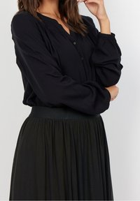 Frau trägt eine schwarze Bluse mit langen Ärmeln und Knöpfen sowie einen schwarzen plissierten Rock, posiert mit einem Arm verschränkt und der Hand nahe dem Gesicht.