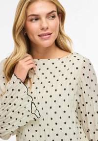 Biała bluzka w czarne kropki, z teksturowanej tkaniny, z falbankami na rękawach. Posiada prosty dekolt i delikatne czarne wykończenie na krawędziach.