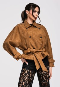 Femme aux cheveux bruns attachés en queue de cheval, portant une veste en daim marron ceinturée et un pantalon noir en dentelle, posant de profil devant un fond uni.