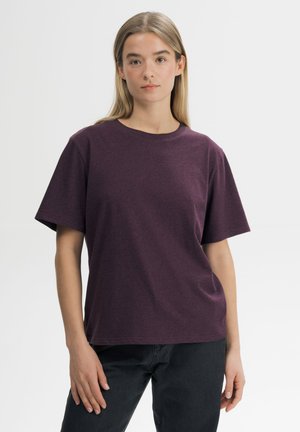 Jonge vrouw met lang blond haar die een effen donkerpaarse korte mouw t-shirt en zwarte broek draagt, staande tegen een witte achtergrond.