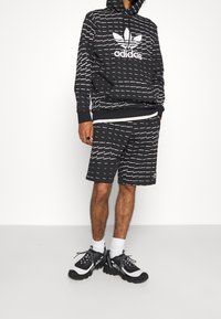 Svart hoodie och shorts set med vitt upprepande logotypmönster. Hoodien har dragsko och känguruficka. Bärs med svarta sneakers.