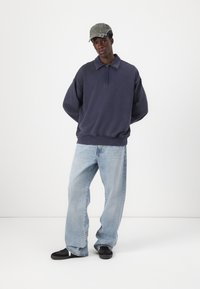 Pullover navy con colletto con zip, vestibilità comoda, abbinato a jeans a gamba larga azzurri e scarpe nere. Un cappellino grigio completa il look.