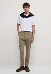 Giovane uomo in piedi, indossa una camicia bianca a maniche corte, un maglione nero drappeggiato sulle spalle, pantaloni beige e stivali in camoscio marrone.