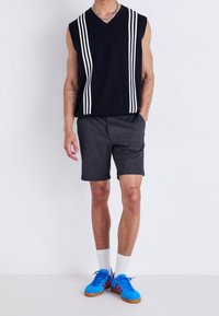 Maglione nero senza maniche con strisce bianche, abbinato a pantaloni corti grigi e scarpe da atletica blu. Indossato con calzini bianchi fino alla caviglia.