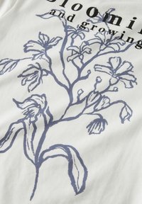 Zweig mit blauen Blumenillustrationen und teilweise schwarzem Text, der "blühen und wachsen" auf weißem Stoff liest.