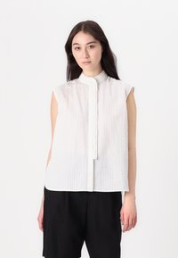 Hvit ärmeløs bluse med høy krage, med strukturert stoff og knappelukking foran, parret med svarte shorts.