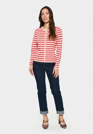 Cardigan a strisce rosse e rosa con chiusura a bottoni, maniche lunghe e polsini a coste, abbinato a jeans blu scuro e scarpe a fantasia.