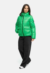 Heldere groene pufferjack met een hoge kraag, ritsluiting en twee voorzakken, gecombineerd met losse zwarte broeken en witte sneakers.