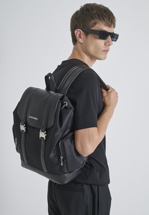 Mochila negra con textura suave, que presenta herrajes plateados y cremalleras laterales dobles. Correas de cuero ajustables y un diseño estructurado.