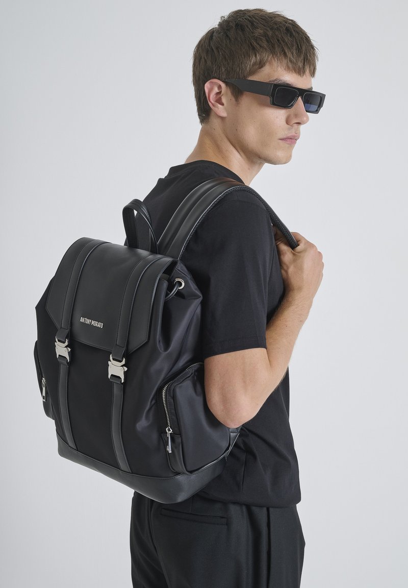 Zaino nero con una texture liscia, dotato di hardware argentato e doppie cerniere laterali. Spallacci in pelle regolabili e design strutturato.