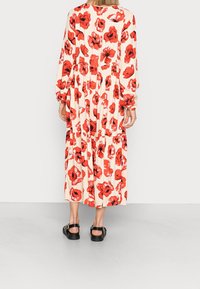 Femme portant une longue robe crème à imprimé floral rouge, avec des manches longues et des sandales noires, se tenant devant un mur uni de couleur claire.