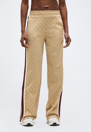 Person wearing beige patterned wide-leg pants with maroon and cream side stripes and white sneakers, standing against plain background.