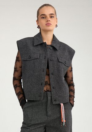 Jeune femme debout portant un gilet structuré gris foncé et un pantalon assorti sur un top en dentelle noire transparente à manches longues, mains dans les poches.