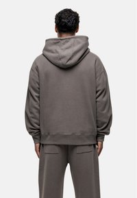 Felpa grigia oversize con cappuccio, polsini e orlo a coste. Il tessuto è morbido, texturizzato, con una vestibilità rilassata. La vista posteriore non mostra alcun marchio visibile.