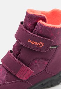 Lila Winterstiefel mit strukturiertem Obermaterial, orangefarbener Fleece-Fütterung, doppelten Klettverschlüssen und gemustertem Design. Schwarze Gummisohle für gute Traktion.