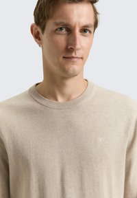 Licht beige gebreide trui met een ronde hals, geribbelde kraag en subtiel logodetail op de borst. Zachte textuur, getailleerd model.