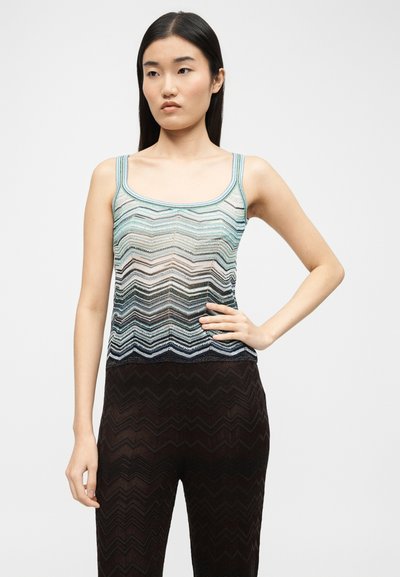 Νέα γυναίκα με μακριά μαύρα μαλλιά, φορώντας ένα αμάνικο πλεκτό τοπ με παστέλ ρίγες και σκούρο παντελόνι με σχέδιο ζιγκ-ζαγκ, στέκεται με ένα χέρι στη μέση.