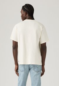 Crème korte mouwen t-shirt van katoen, met een ontspannen pasvorm en een label aan de onderlinkerkant. G gedragen met lichtblauwe denim jeans.